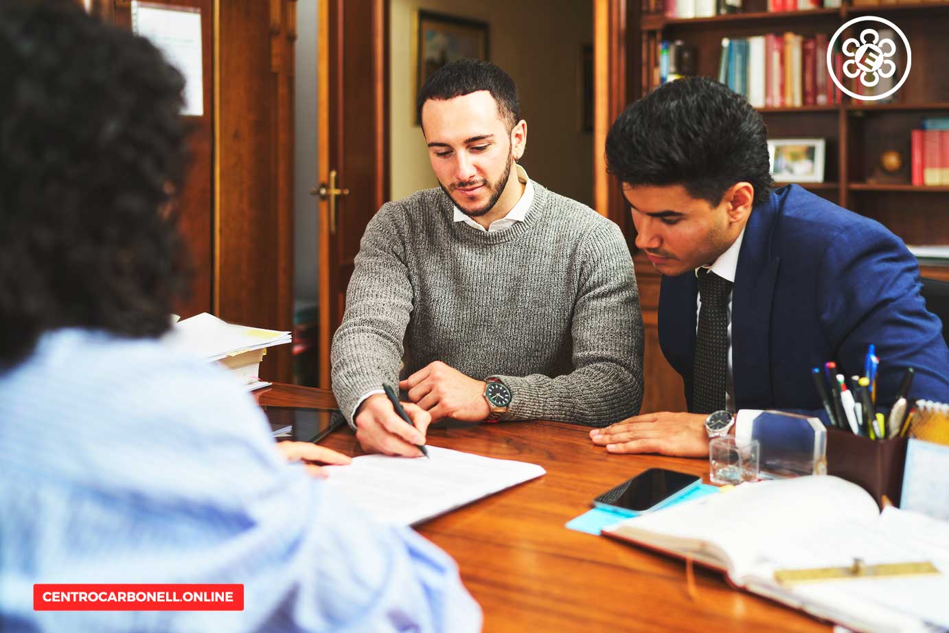 Tres personas discutiendo y tomando notas en una reunión en una oficina, con documentos y materiales de trabajo sobre la mesa.