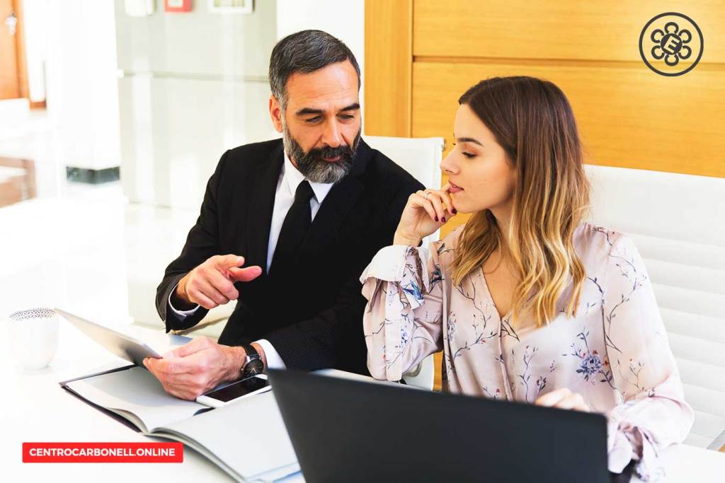 Un hombre de cabello canoso y barba discute con una mujer joven en una oficina, revisando documentos y trabajando en una computadora portátil.