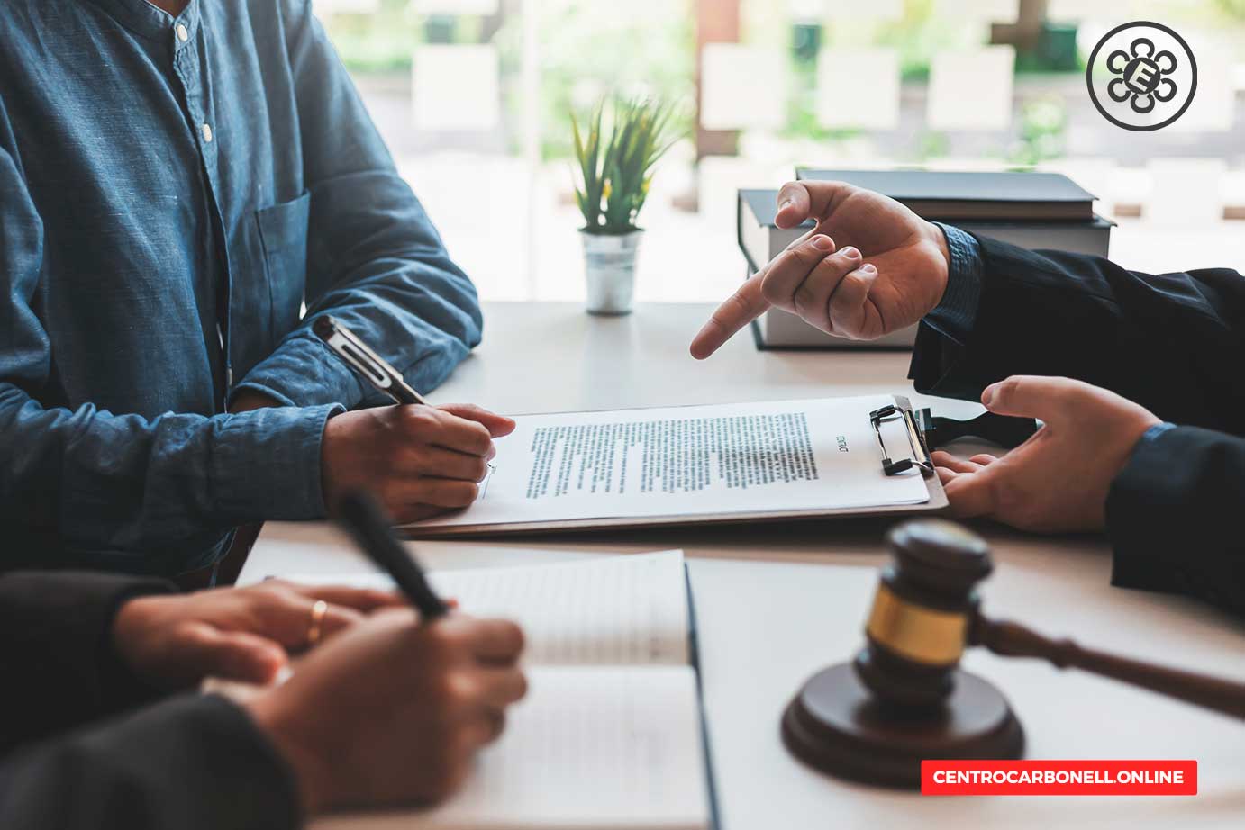 Dos personas discutiendo un documento legal en una mesa, con un mazo de juez y una planta decorativa en el fondo.