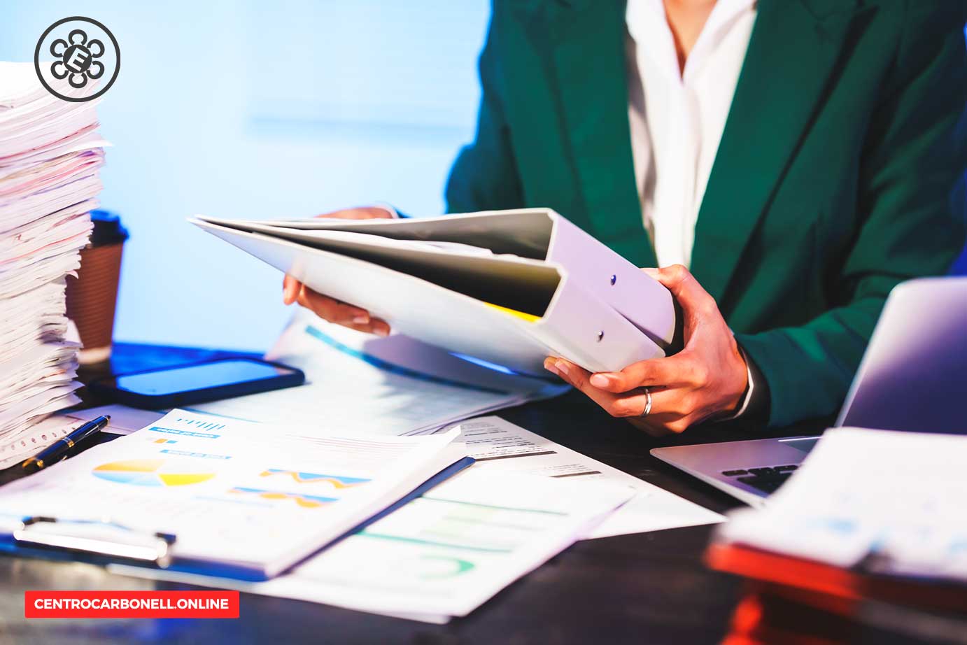 Persona sosteniendo una carpeta con documentos, rodeada de papeles y gráficos en una mesa de trabajo.