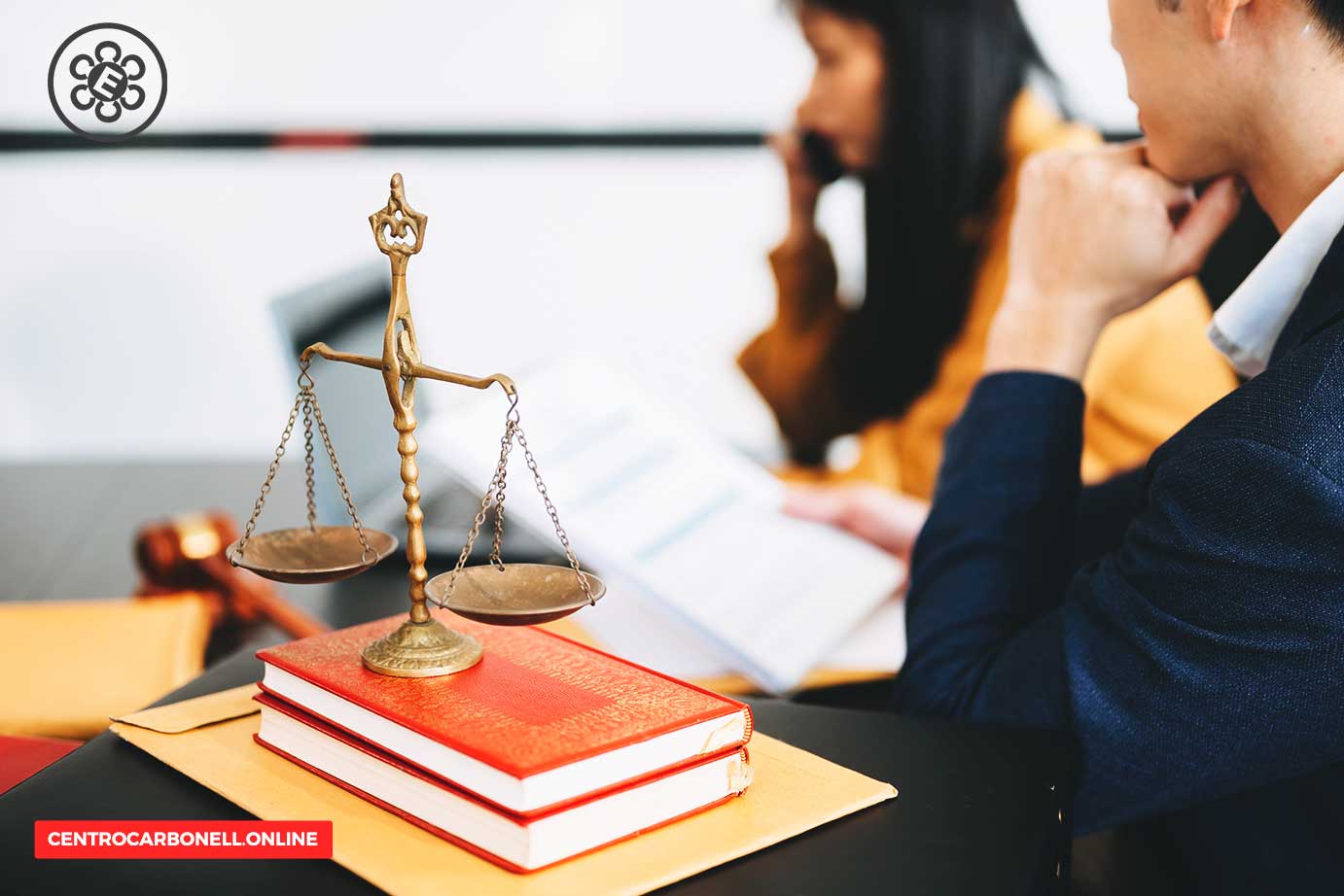 Una balanza de la justicia sobre libros de derecho, con personas en segundo plano en un entorno de discusión legal.