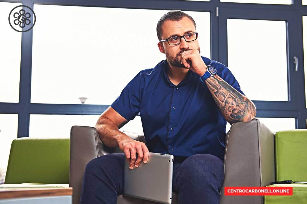 Un hombre sentado en un sofá, sosteniendo una tablet y con expresión pensativa, viste una camisa azul y tiene un tatuaje visible en el brazo.