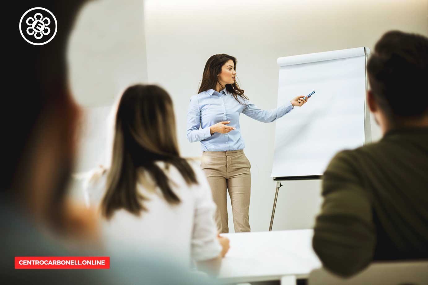 Una presentadora en una sala de capacitación muestra información en una pizarra blanca mientras la audiencia observa desde su asiento.