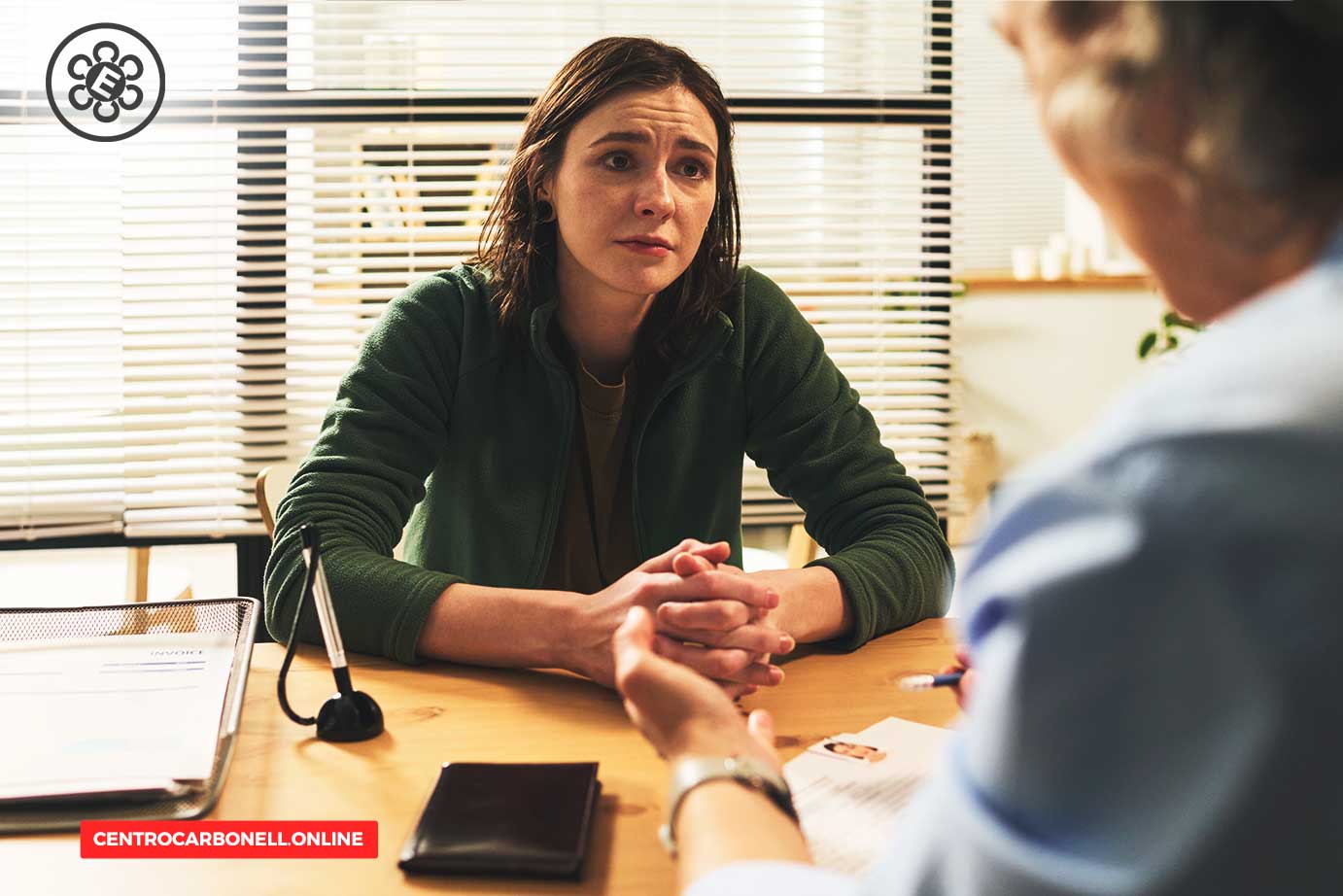 Una mujer sentada frente a otra persona en una mesa, mostrando expresión de preocupación durante una conversación, en un entorno de oficina con ventanas con persianas.