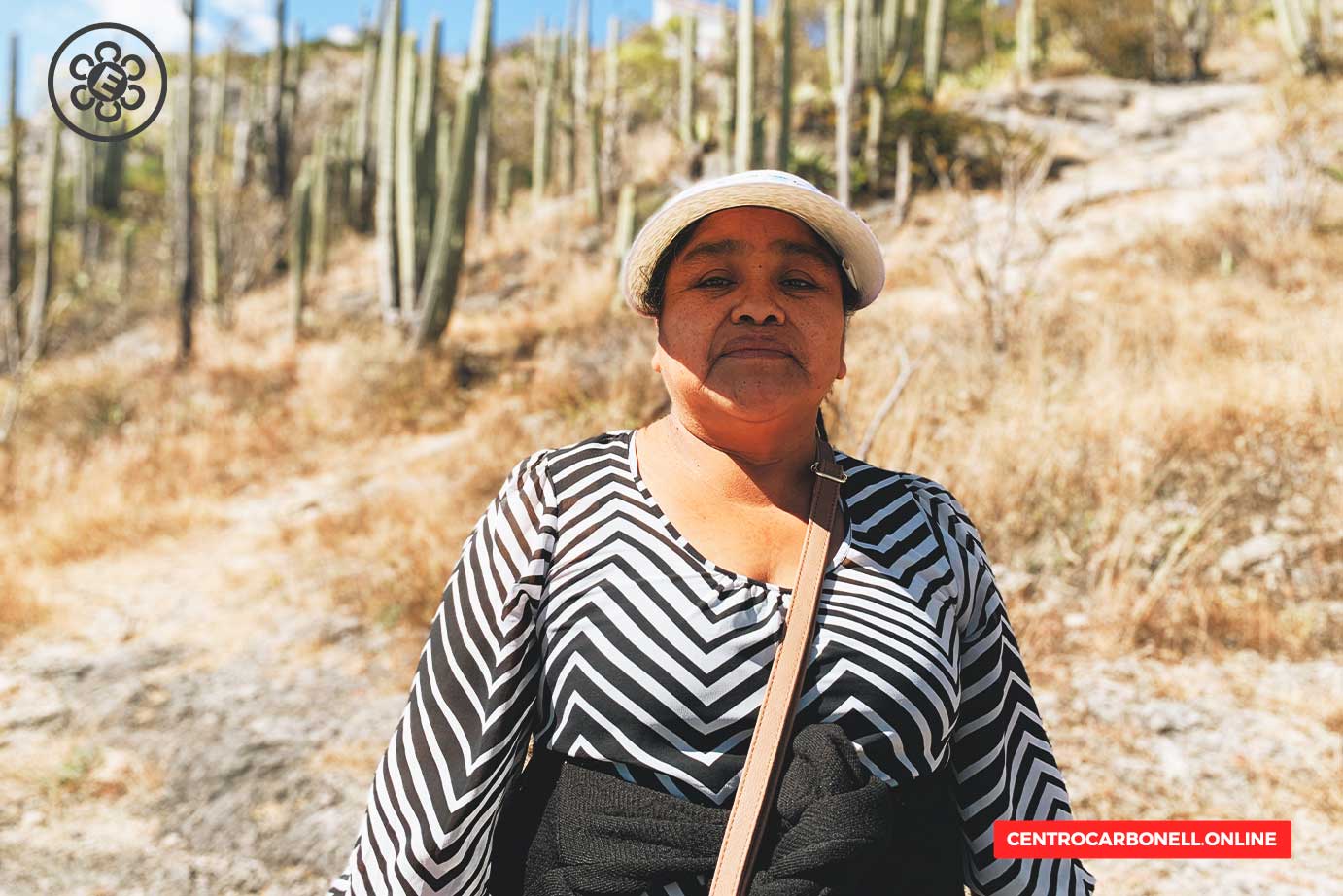 Una mujer de pie frente a un paisaje seco con cactáceas, vistiendo una blusa de rayas en blanco y negro y un sombrero. La escena captura un entorno natural con un cielo despejado.