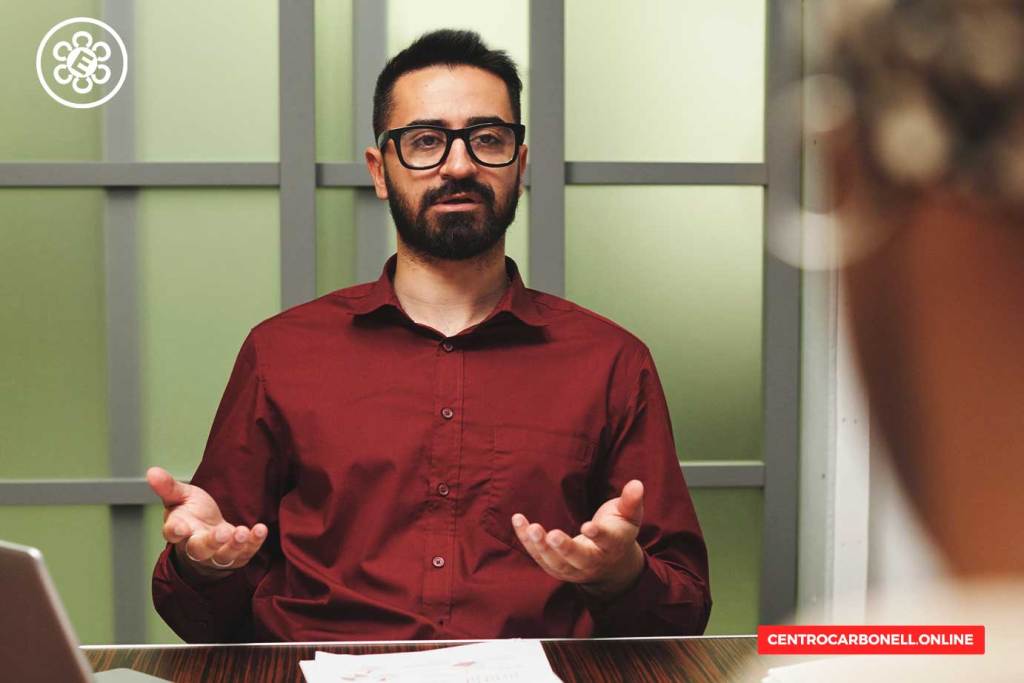 Hombre con gafas de vista, barba y camisa roja gesticula mientras habla en una reunión o entrevista, con una expresión seria y reflexiva.