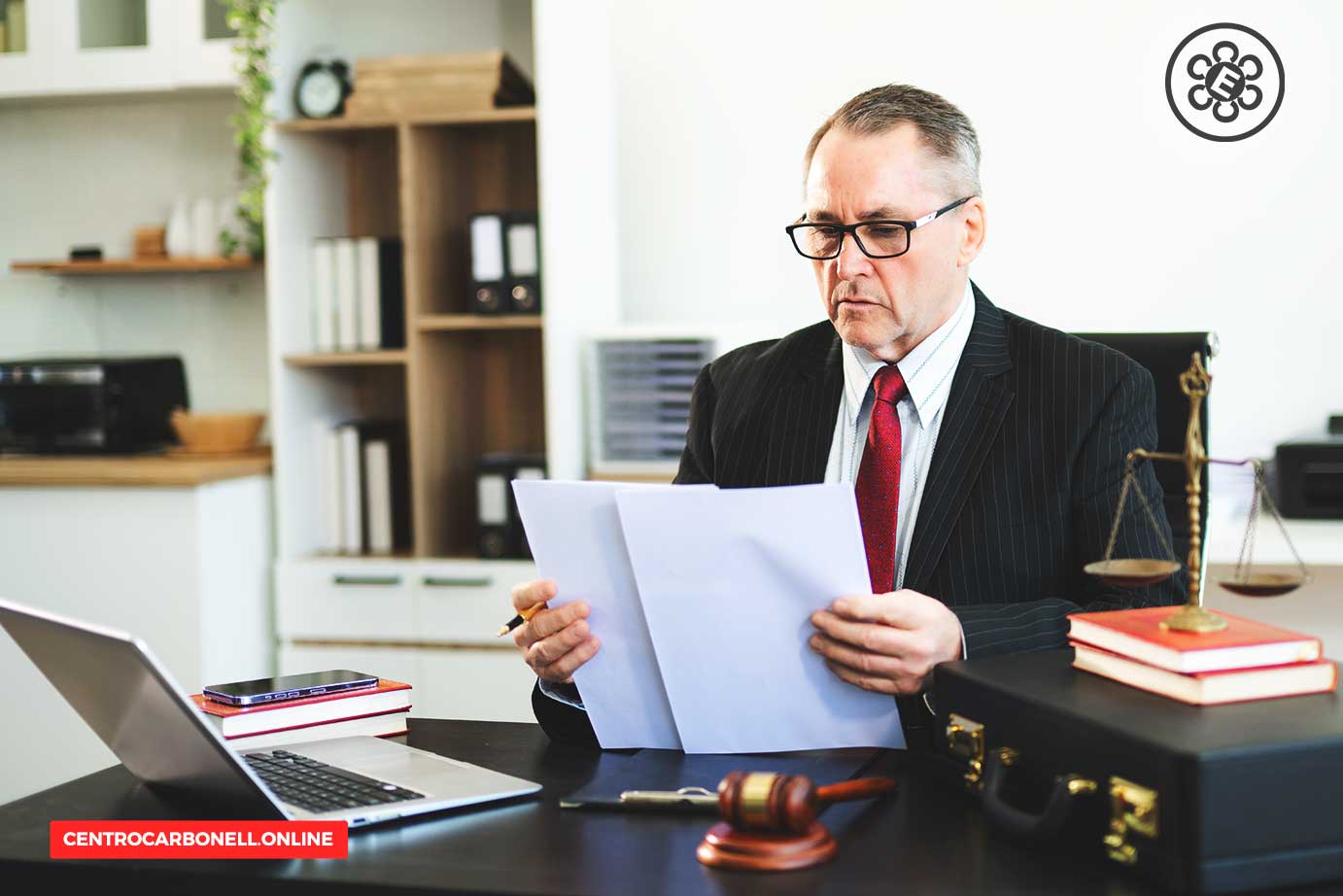Abogado sentado en un escritorio revisando documentos, con una computadora portátil y una balanza de justicia en el fondo.