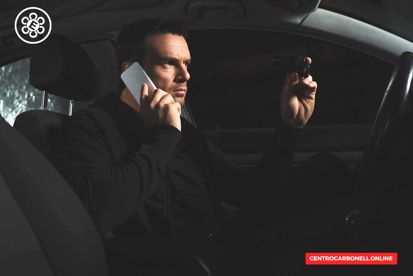 Hombre sentado en un automóvil, utilizando un teléfono móvil con una mano y sosteniendo un dispositivo de grabación con la otra, mirando hacia adelante con expresión seria.