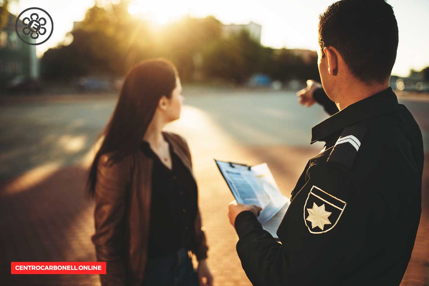 Un oficial de policía sostiene un documento mientras habla con una mujer en un entorno urbano iluminado por la luz del sol.