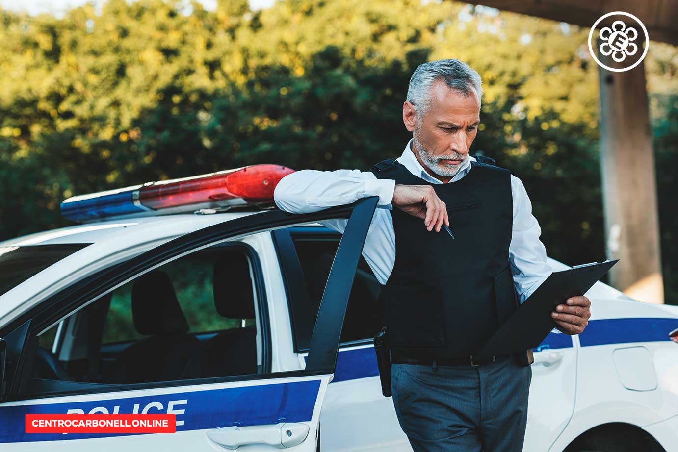 Un oficial de policía revisando un informe junto a un coche policial con luces encendidas, en un entorno al aire libre.