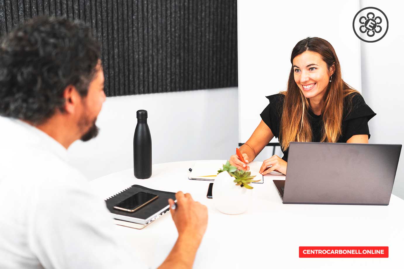 Dos profesionales en un entorno de trabajo colaborativo, sentados en una mesa con un computador portátil, una botella de agua y material de oficina, durante una conversación animada.
