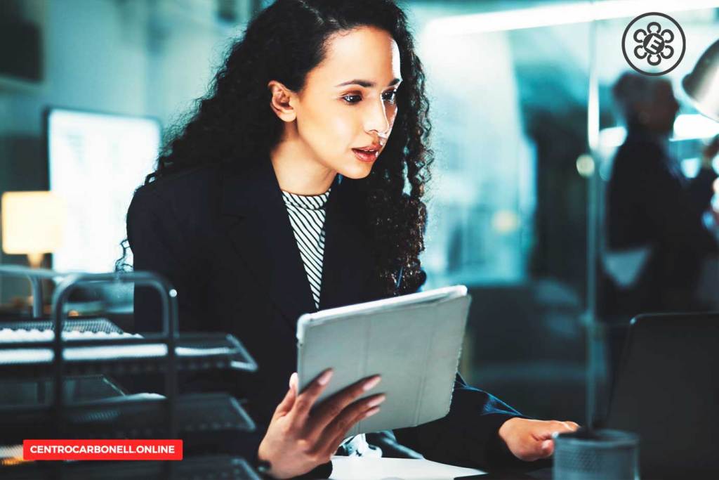 Una profesional con cabello rizado, vestida con un blazer, observa detenidamente una tablet en un entorno laboral moderno, con computadoras y otros elementos de oficina en el fondo.