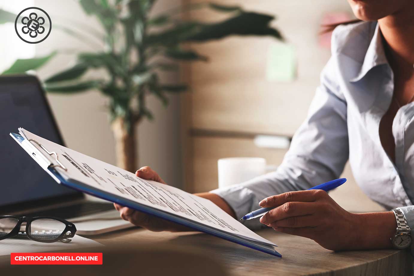 Una persona revisando un documento en un portafolio mientras sostiene un bolígrafo, con una computadora portátil y unas gafas visibles en la mesa.