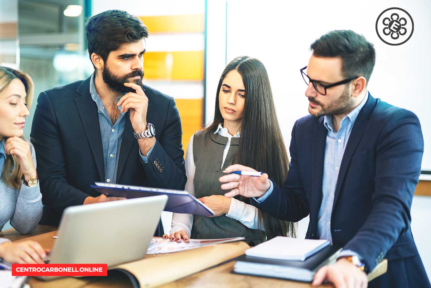 Un grupo de cuatro profesionales discutiendo y analizando documentos en una mesa, con una laptop y herramientas de oficina visibles.