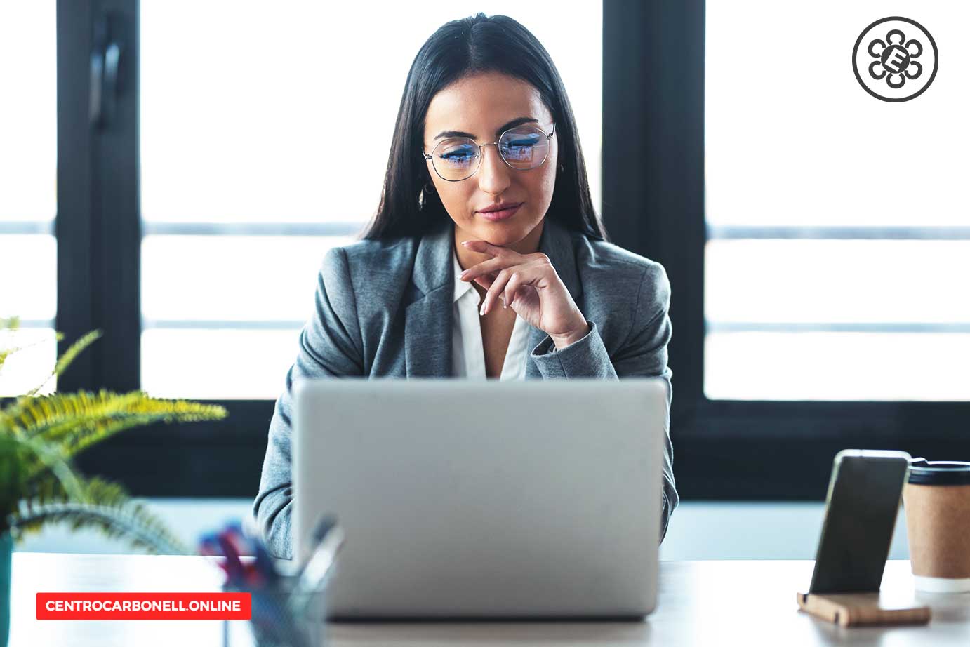 Mujer joven trabajando en una computadora portátil en una oficina moderna, con una planta y una taza de café en la mesa.