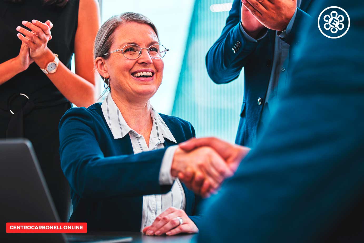 Una mujer sonriente estrechando la mano de un hombre en un entorno de oficina, con otros adultos aplaudiendo en el fondo.