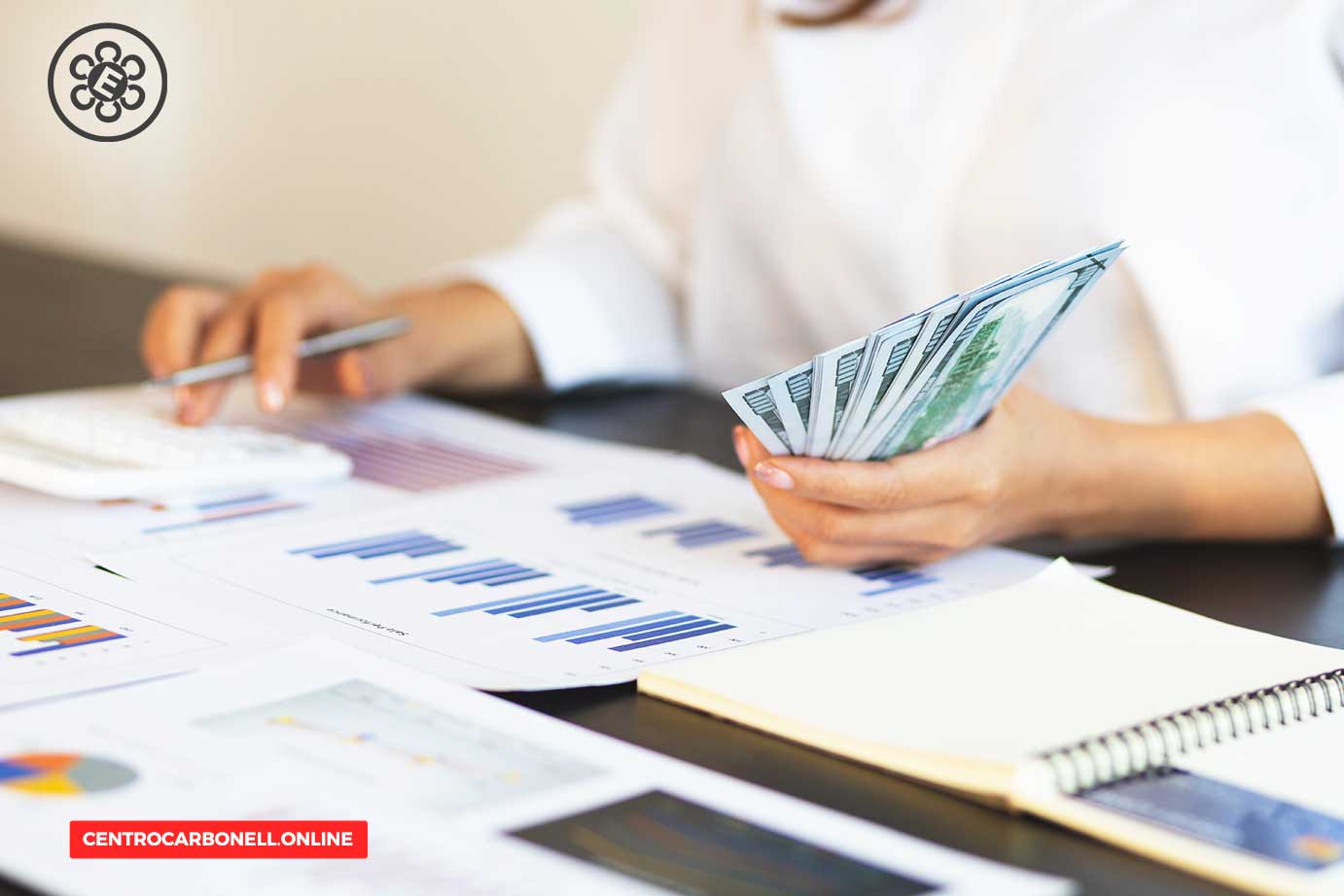 Una persona sosteniendo billetes de dólares mientras utiliza una calculadora y revisa documentos con gráficos en una mesa de trabajo.