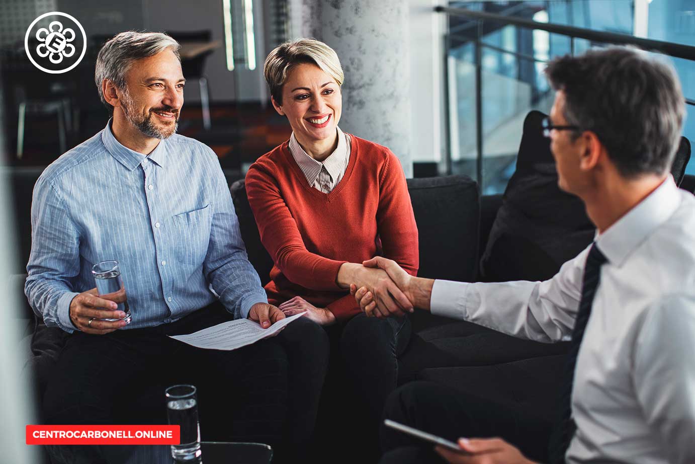 Tres personas en una reunión, una mujer y un hombre sonrientes sosteniendo documentos, mientras un tercer hombre les estrecha la mano.