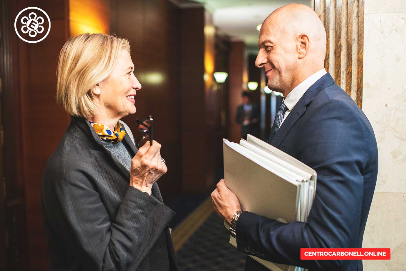 Una mujer y un hombre conversan en un ambiente profesional, sonriendo. La mujer sostiene unas gafas y el hombre lleva varios documentos en sus manos.
