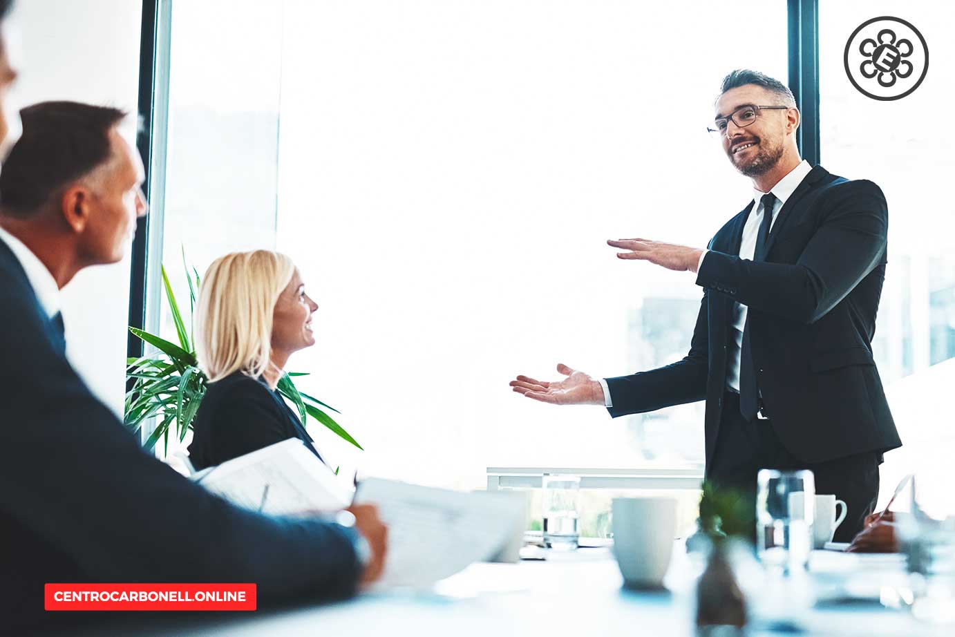 Un abogado en traje negro presenta un tema durante una reunión, mientras sus colegas lo observan atentamente en una sala iluminada con luz natural.