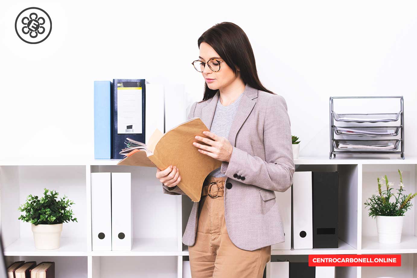 Una mujer de cabello largo y castaño, vestida con un blazer claro, sostiene un expediente en sus manos mientras revisa documentos en una oficina moderna, con estanterías blancas y plantas decorativas al fondo.
