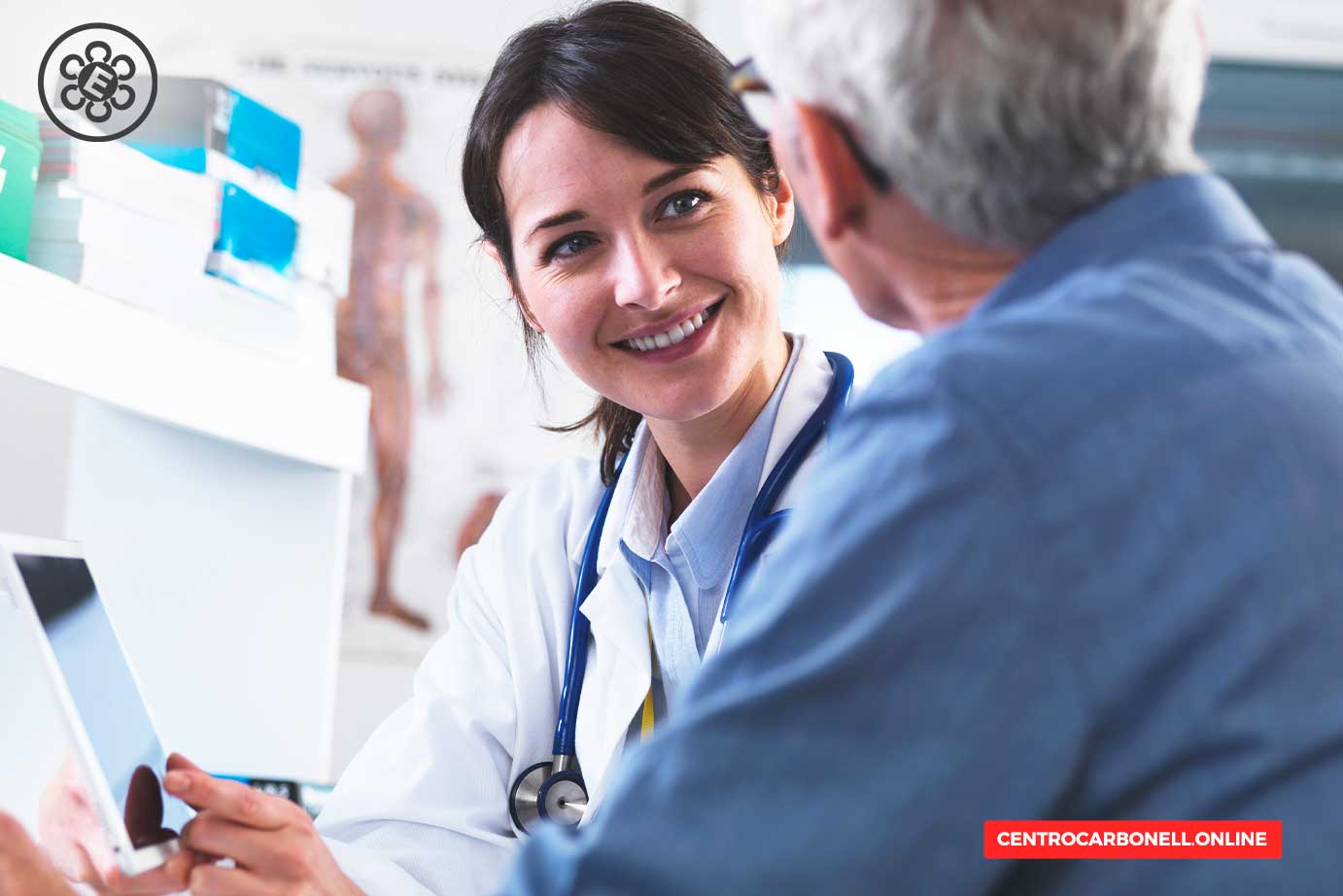 Una doctora sonriente conversando con un paciente durante una consulta médica, con materiales médicos y un gráfico anatómico en el fondo.