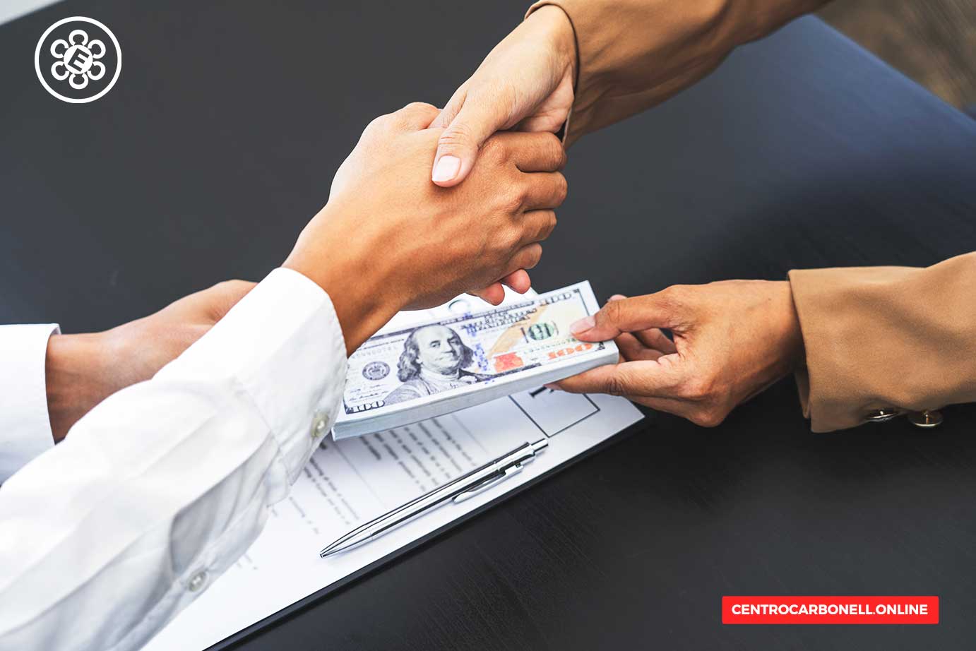 Dos manos estrechándose mientras se entrega un fajo de billetes de dólar sobre una mesa con documentos y un bolígrafo.