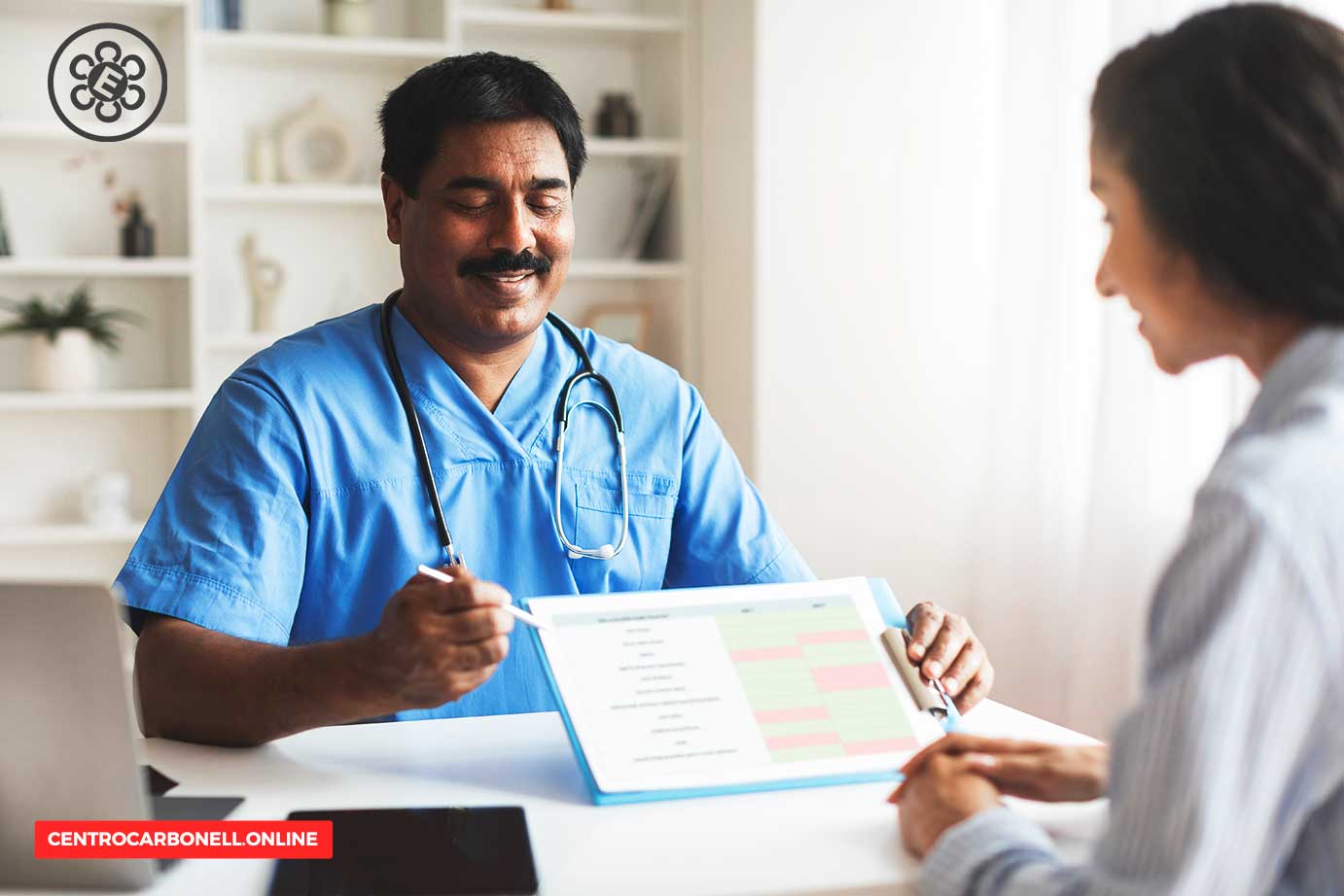 Médico en bata azul sonriendo mientras revisa un documento con una paciente en una consulta médica.