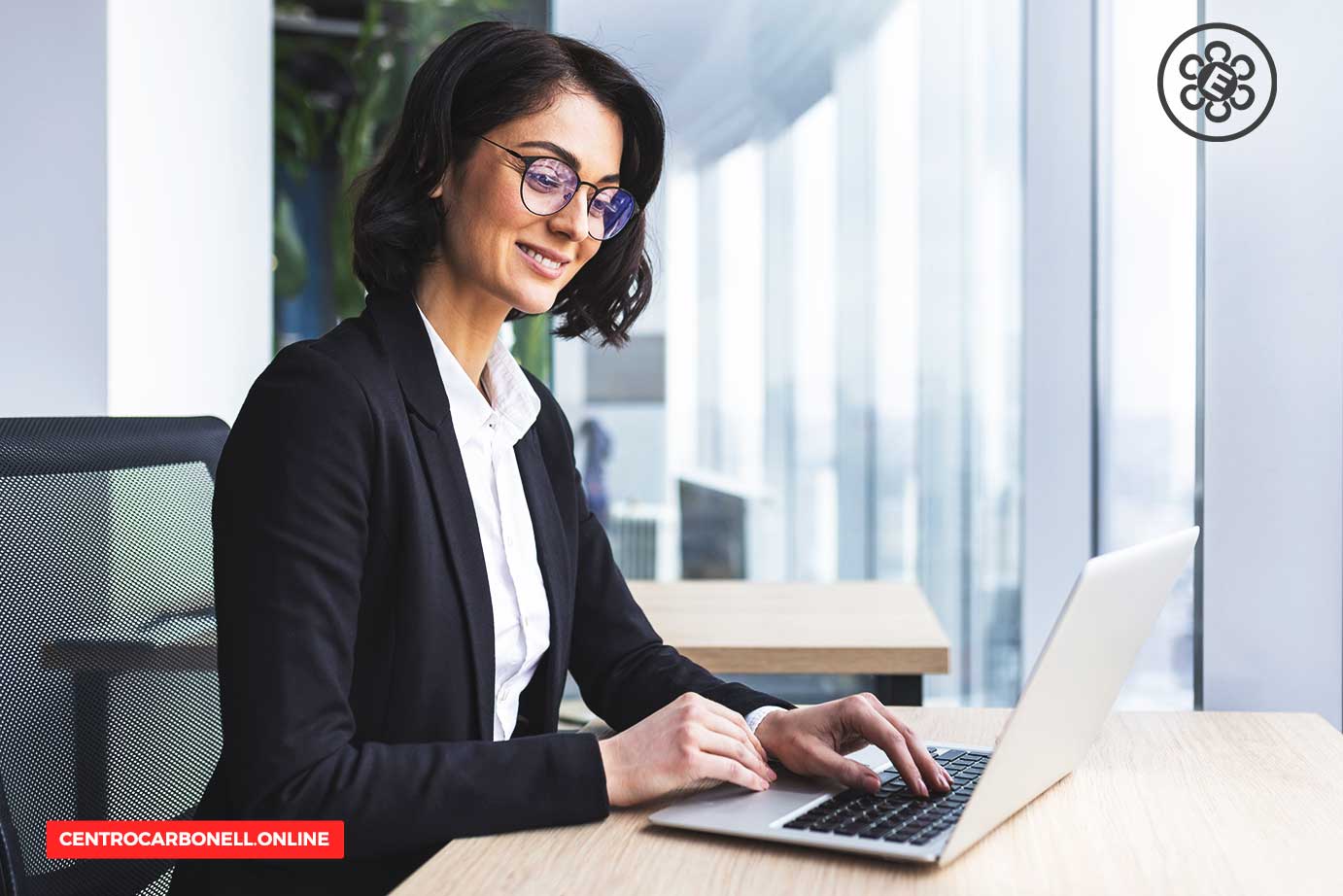 Mujer sonriente trabajando en una computadora portátil en una oficina moderna, vestida con un traje profesional.