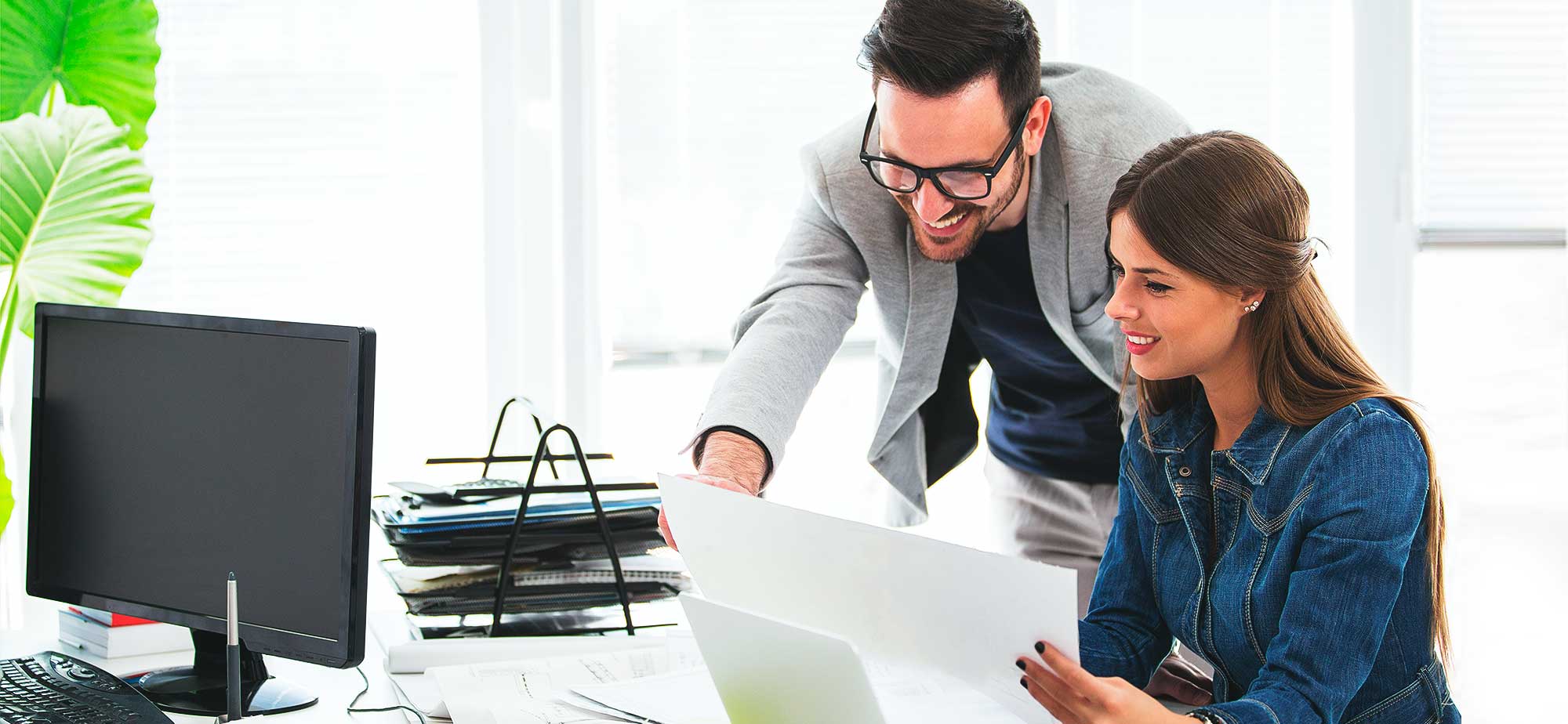 Dos personas en una oficina colaborando sobre documentos, con una computadora y documentos en el escritorio.