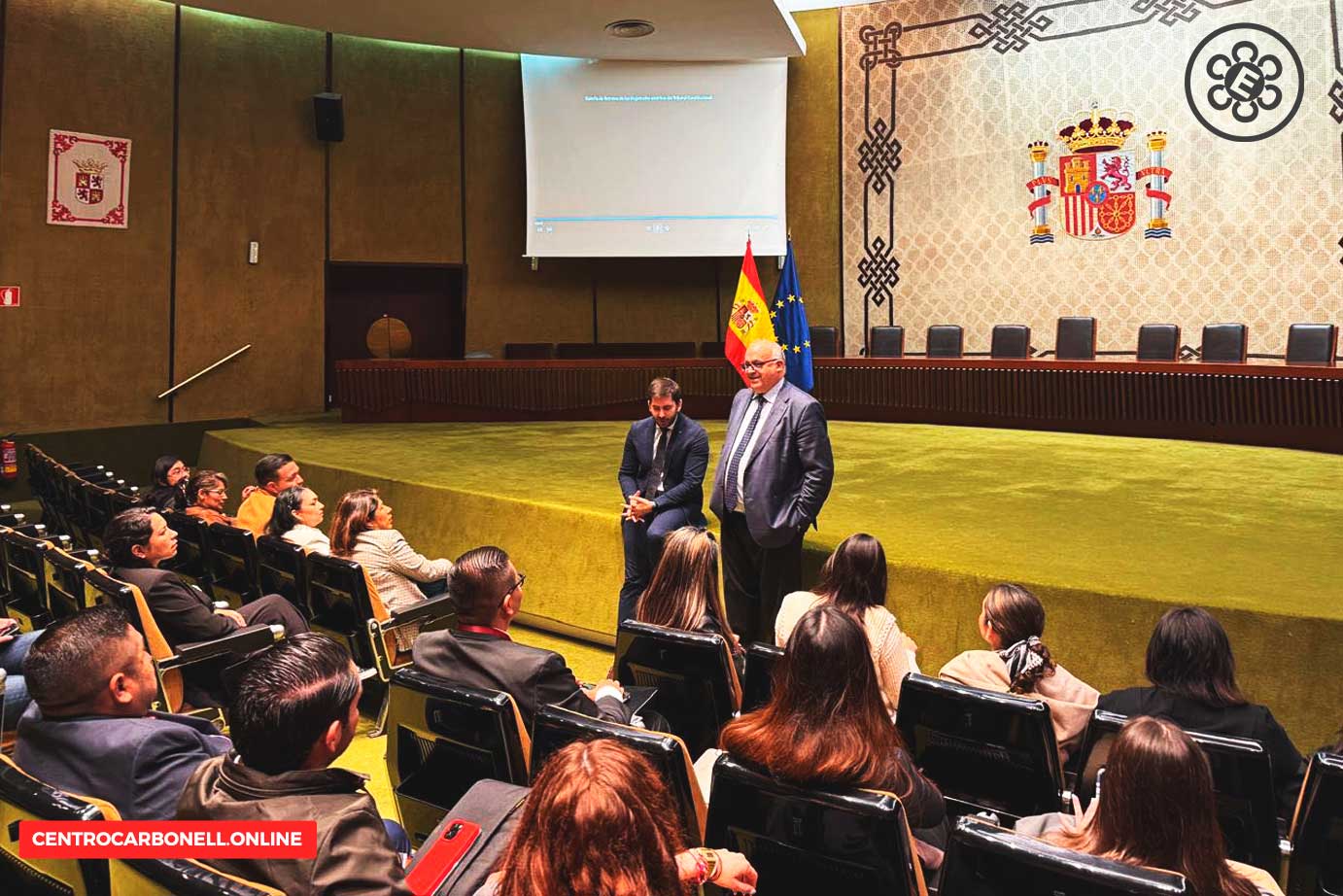 Estudiantes y ponentes durante una conferencia en una sala académica en España, con banderas de España y la UE de fondo.