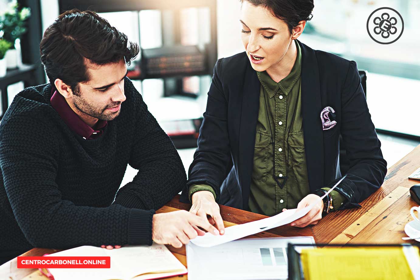 Dos personas revisando documentos en una mesa de trabajo, una de ellas señala un papel mientras la otra observa atentamente.