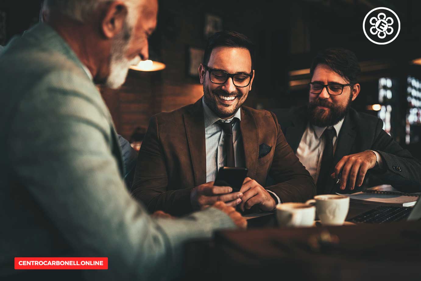 Tres profesionales en un entorno de trabajo informal comparten ideas mientras revisan un teléfono móvil. El ambiente sugiere una conversación amena y colaborativa.