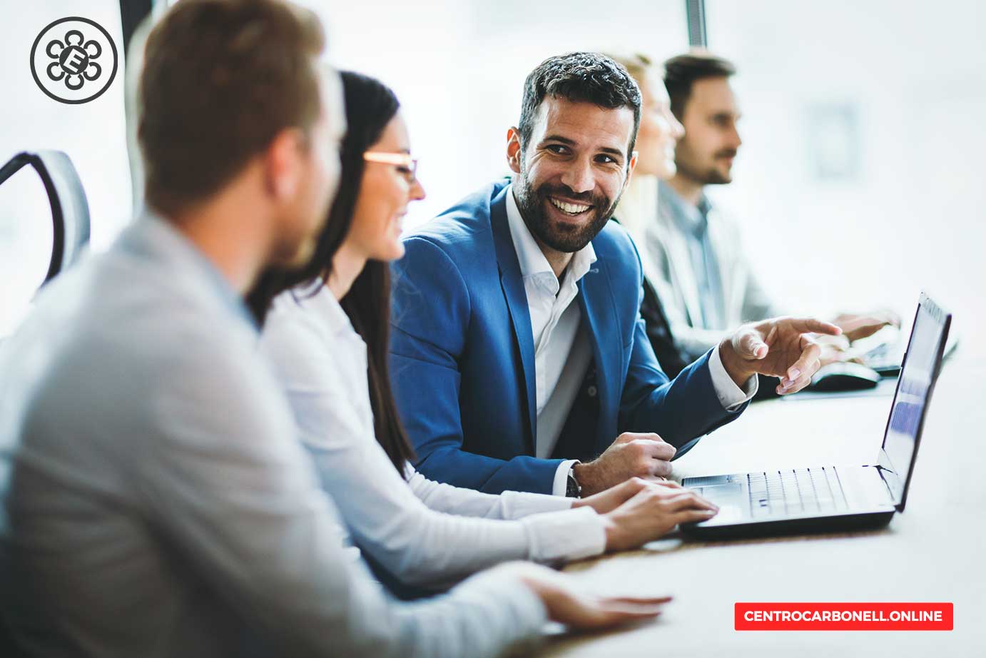 Un grupo de profesionales en una reunión empresarial, discutiendo frente a una computadora portátil. Uno de los hombres está sonriendo y gesticulando mientras habla.