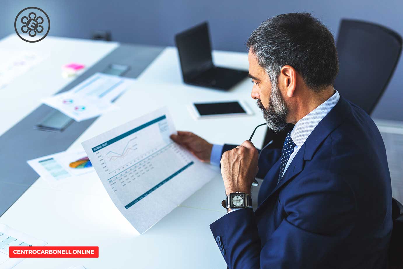 Hombre de negocios analizando un informe en una mesa de trabajo con gráficos y documentos, vistiendo un traje y lentes.