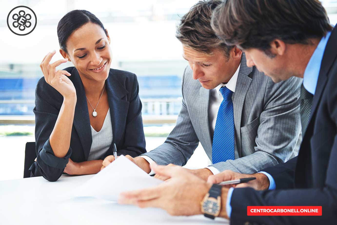 Tres profesionales de negocios revisando documentos en una mesa, todos en trajes formales, con un ambiente de trabajo colaborativo.