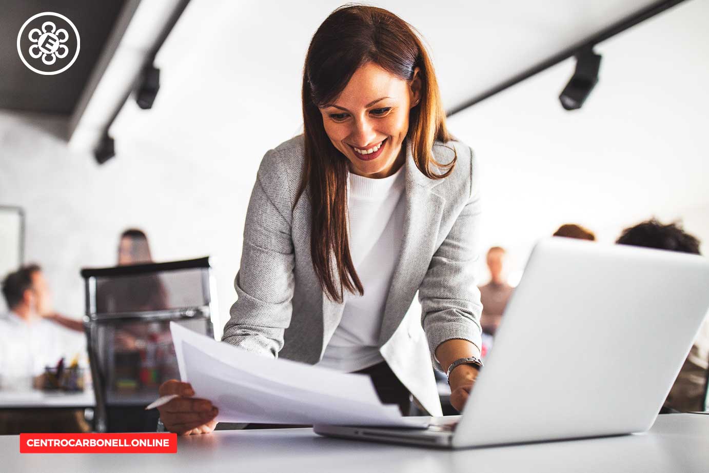 Una mujer sonriente revisando documentos mientras trabaja en una laptop en un ambiente de oficina.