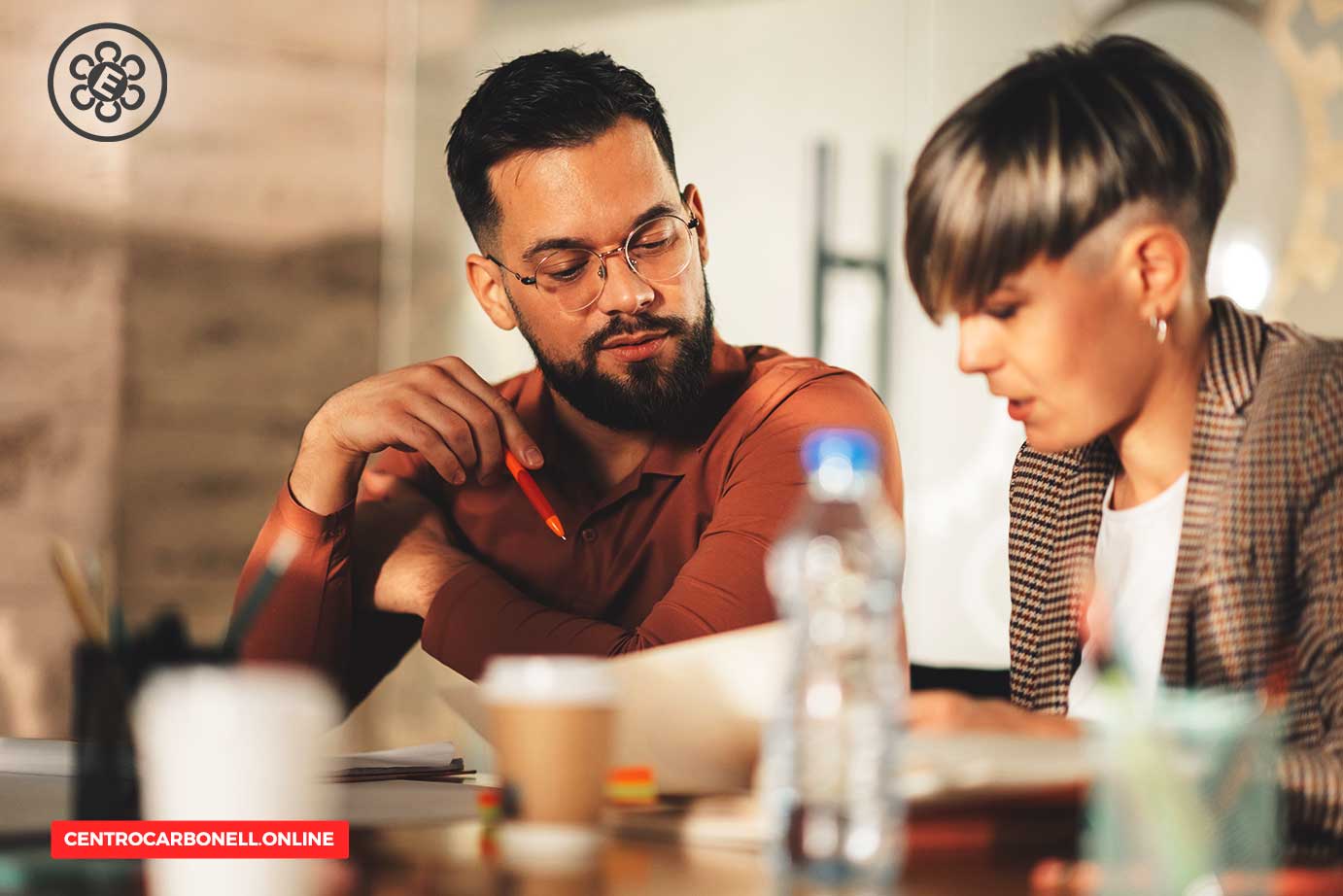 Dos personas sentadas en una mesa, una de ellas revisando documentos mientras la otra observa atentamente, en un ambiente de trabajo colaborativo.