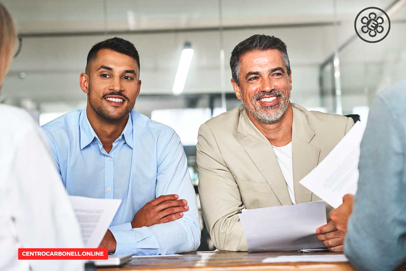 Dos hombres sonrientes sentados en una mesa de reunión, revisando documentos y discutendo en un ambiente de oficina.