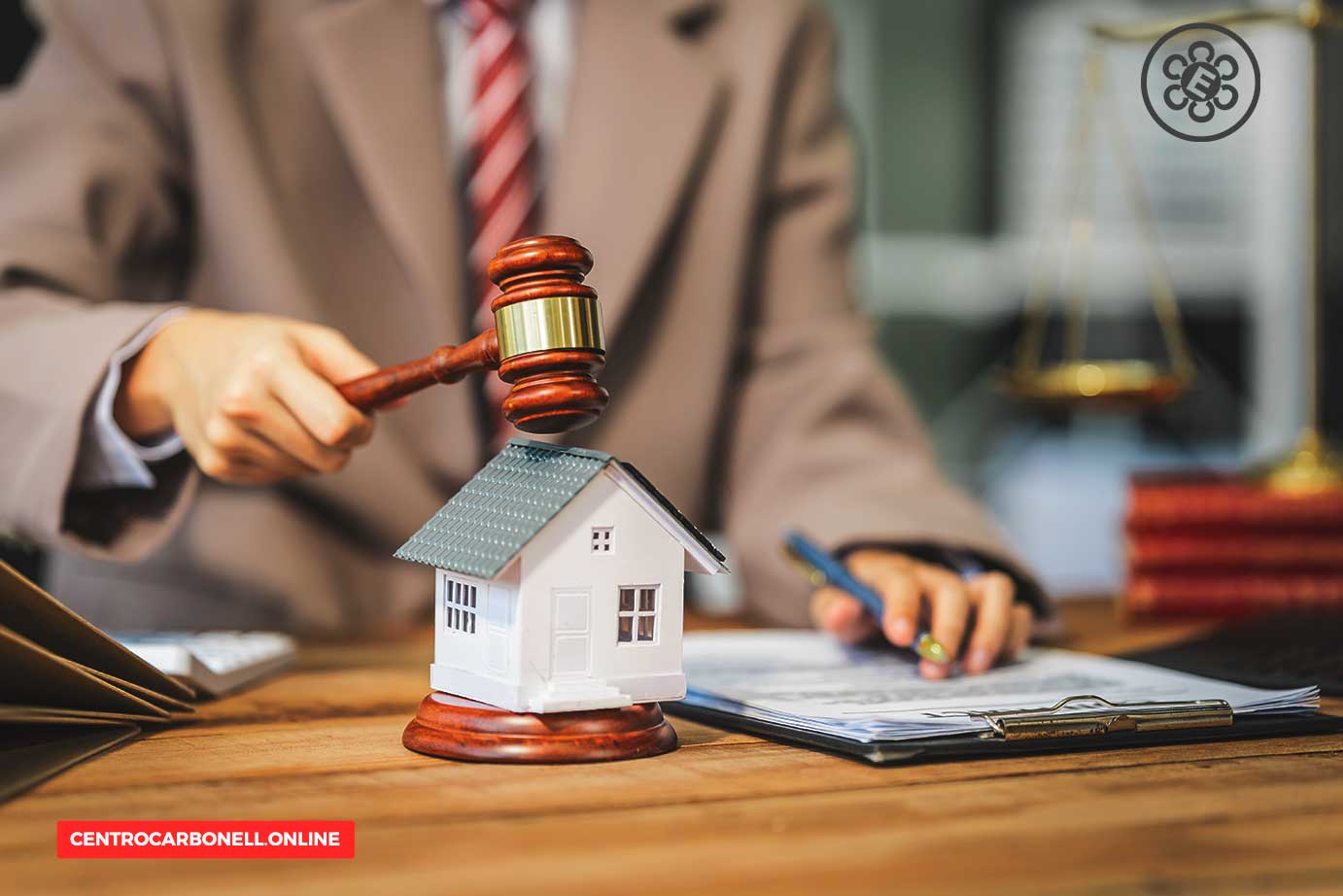 Un juez sostiene un mazo sobre una casa en miniatura, simbolizando decisiones legales relacionadas con bienes raíces, con documentos y balanza en el fondo.
