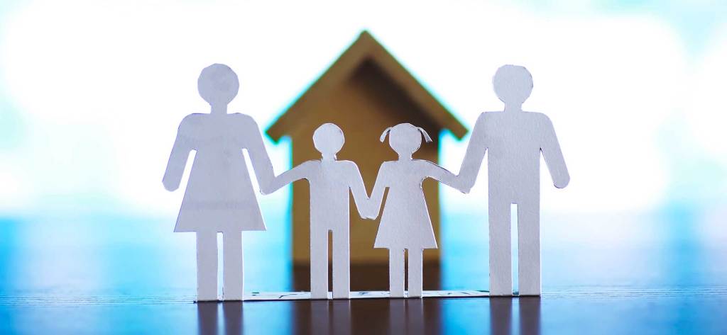 Figuras de papel que representan a una familia, sosteniéndose de la mano, con una casa de fondo difuso.
