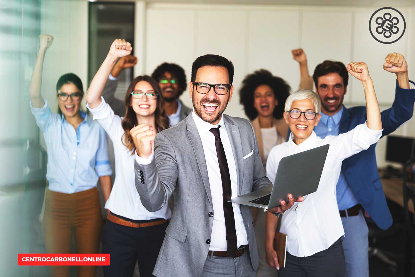 Grupo de personas en una oficina celebrando, con un hombre en traje sosteniendo una laptop y sonriendo, mientras los demás levantan los brazos en señal de victoria.