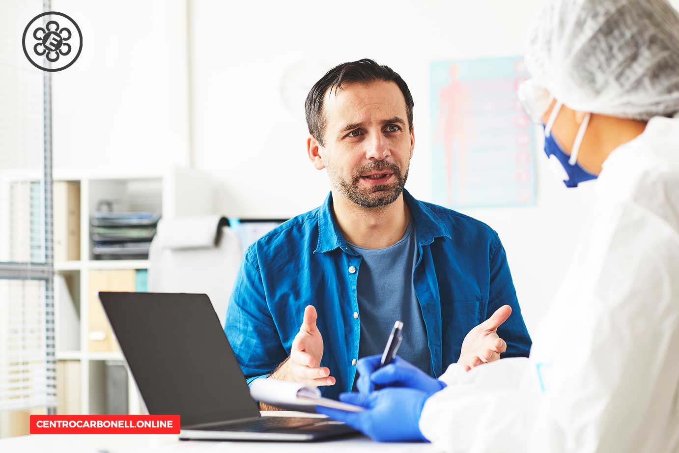 Un médico con equipo de protección escucha atentamente a un paciente, quien parece expresar preocupación. En la mesa hay una computadora portátil y documentos.