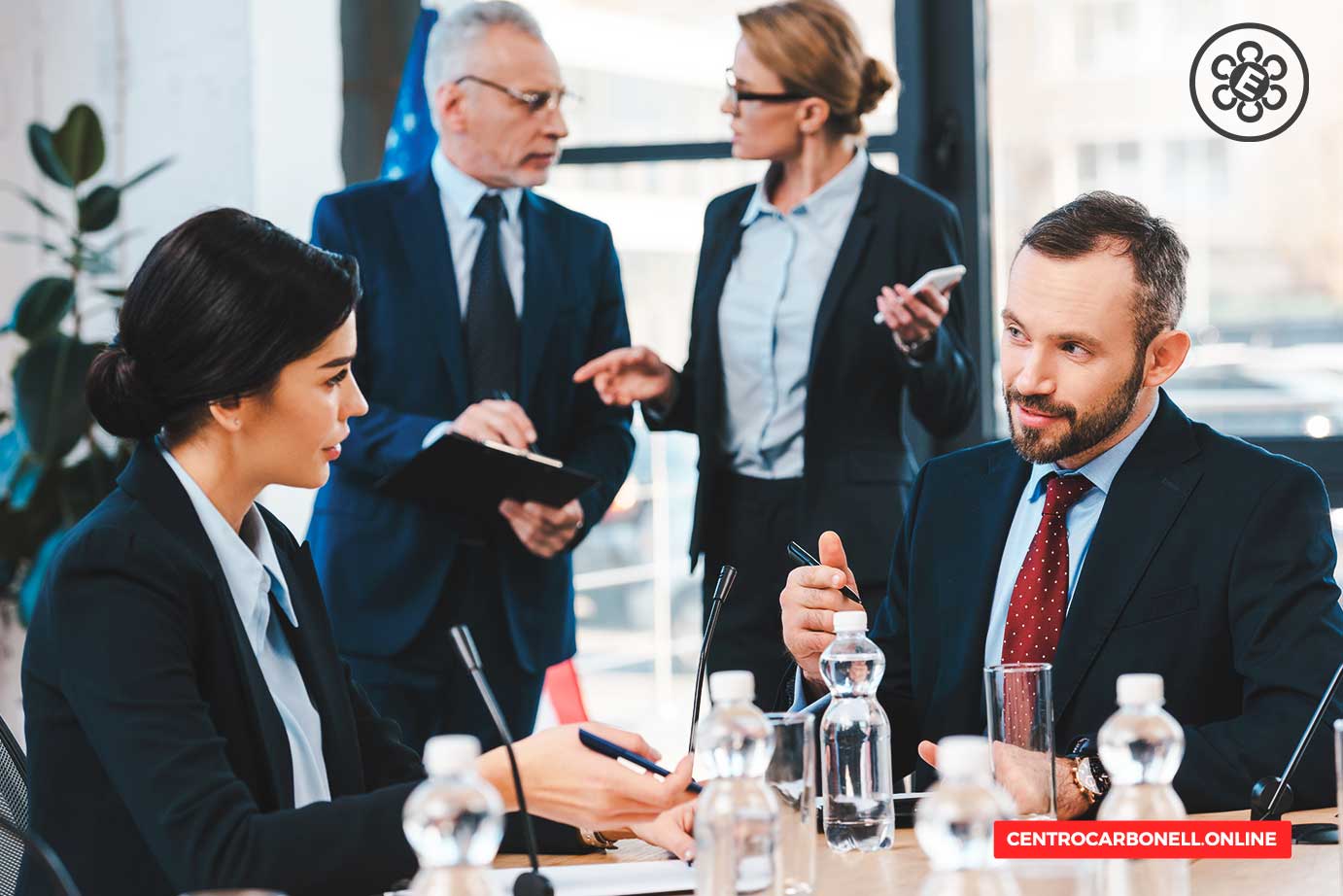 Reunión profesional de abogados en una sala de conferencias, con dos abogados conversando y otros profesionales en el fondo.