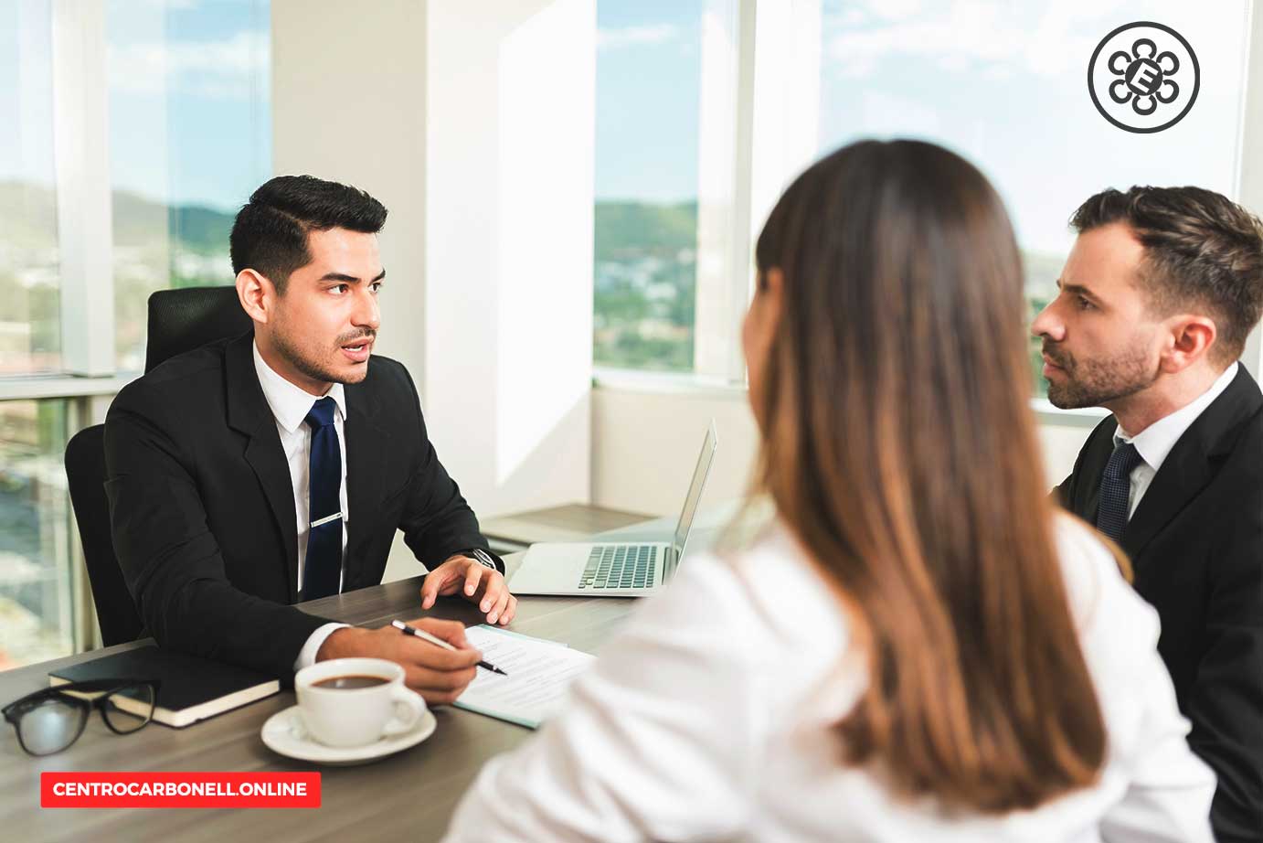 Reunión de negocios en una oficina moderna con tres profesionales discutiendo. Un hombre presenta información mientras dos colegas lo escuchan atentamente.