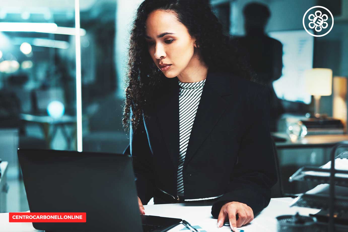 Mujer sentada frente a una computadora portátil en un ambiente de oficina, con una mirada concentrada.