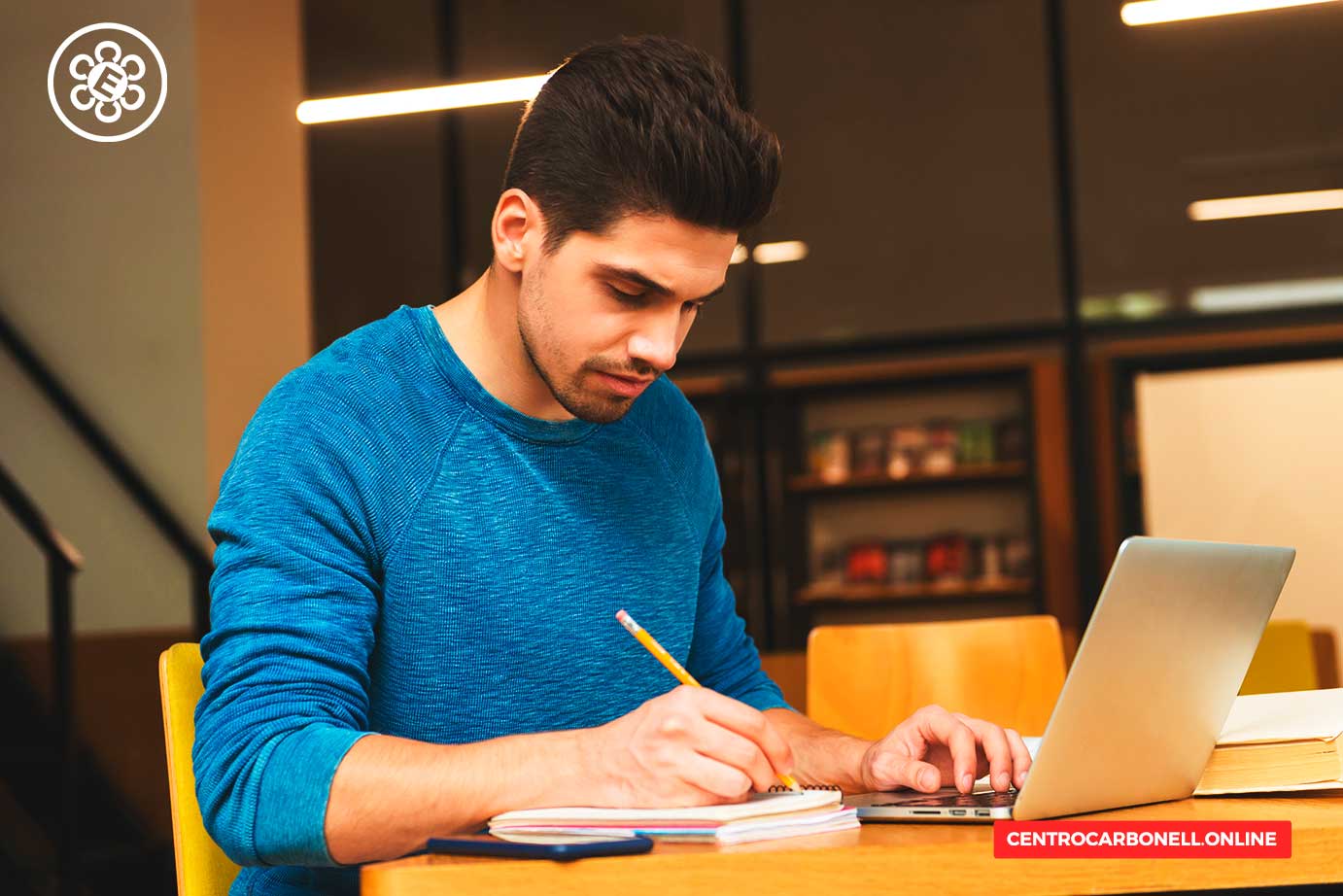 Joven estudiando en una mesa, escribiendo en un cuaderno mientras utiliza una laptop.
