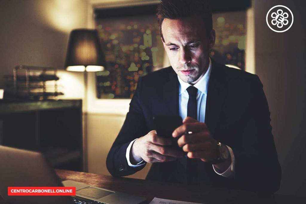 Hombre de negocios sentado en una oficina oscura, mirando su teléfono móvil con atención. Se observan luces de la ciudad a través de la ventana.