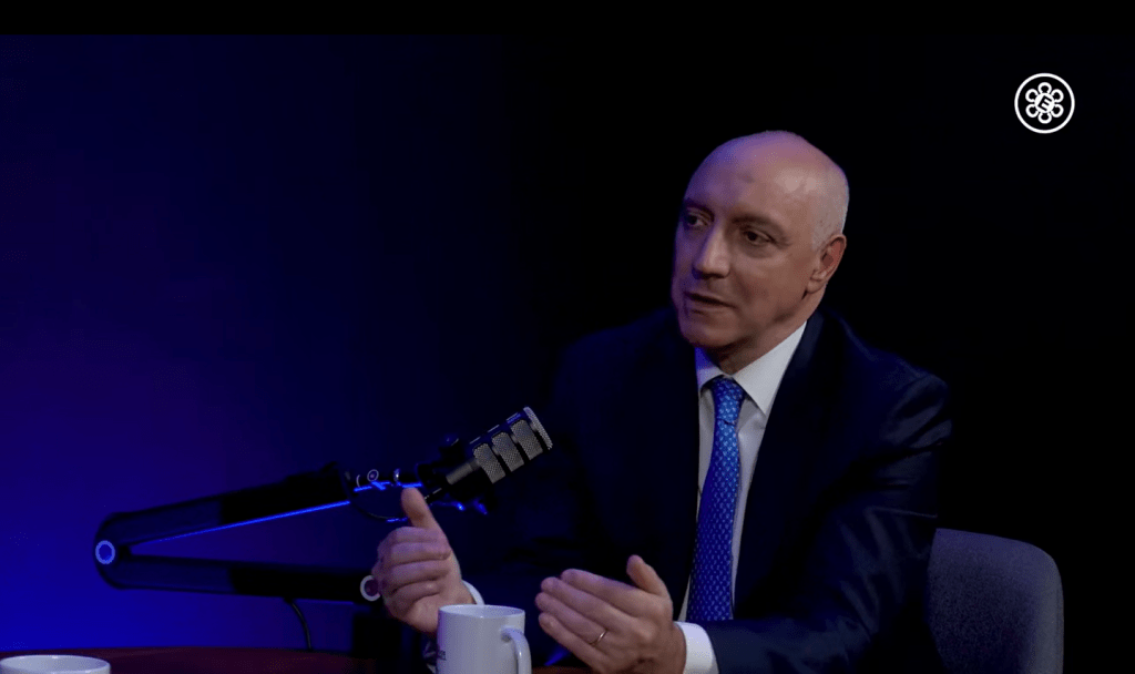 Entrevista a un hombre de negocios con traje en un estudio, hablando frente a un micrófono y una taza.