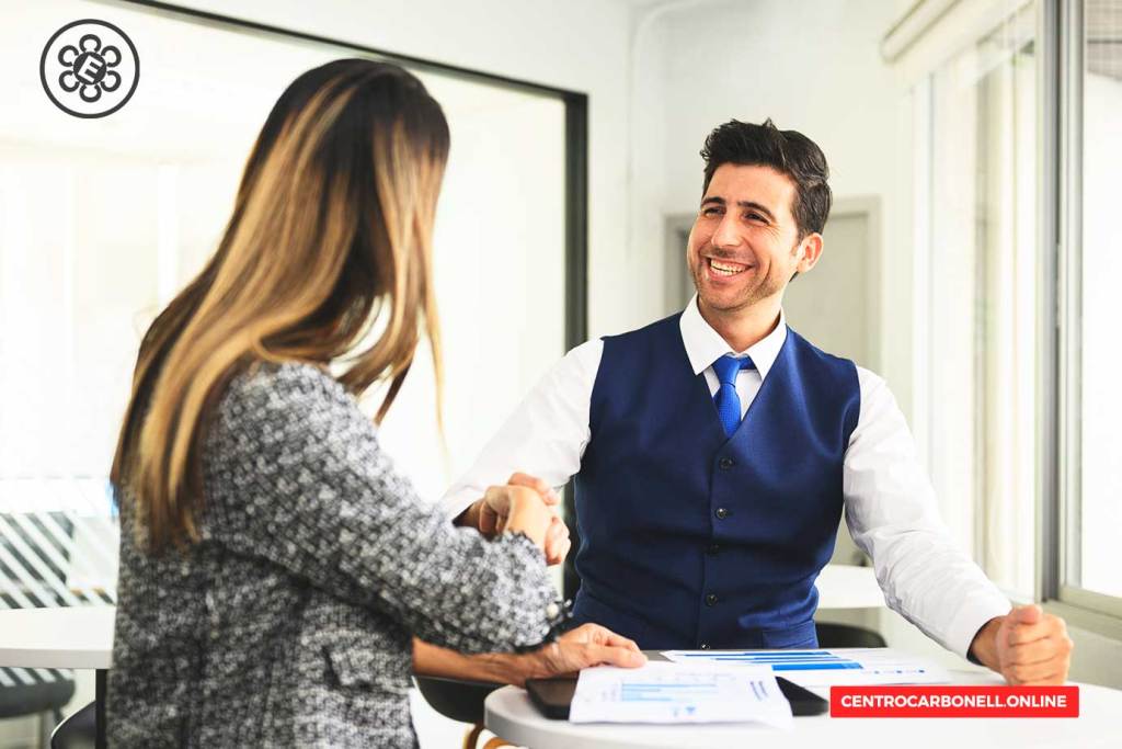 Un hombre sonriente con chaleco azul realiza un apretón de manos con una mujer de cabello largo en un ambiente de oficina moderno. En la mesa hay documentos y gráficos.